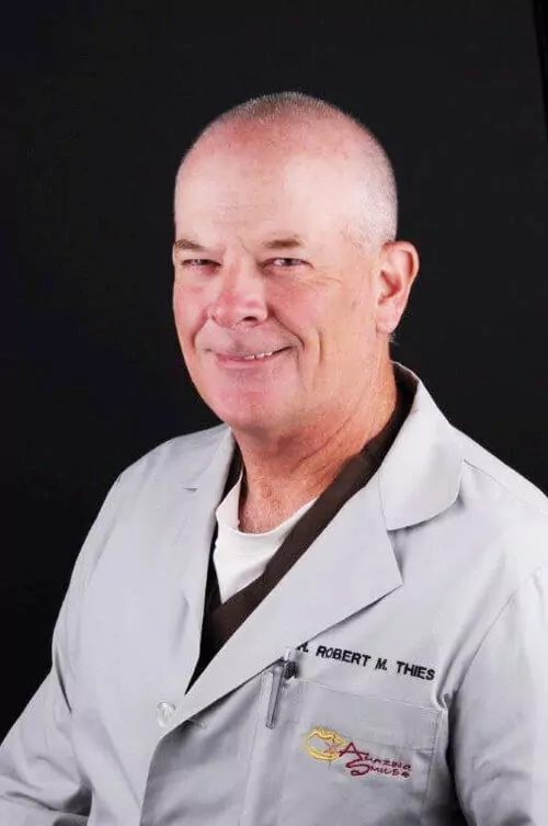 The image shows a person wearing a light gray lab coat with the name "Robert M. Thies" embroidered on it. The lab coat also features a logo with the text "Amazing Smiles" on the left side. The background is black.