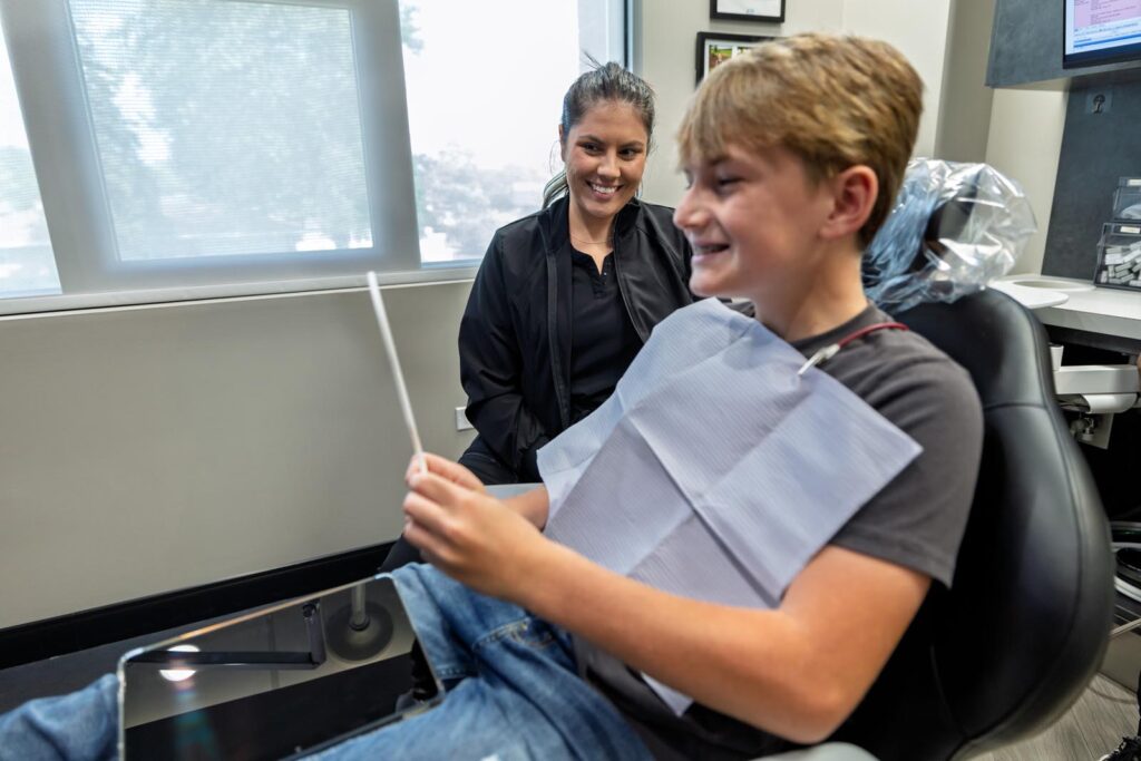 Patient is sitting in a dental chair, holding a mirror, and smiling as he looks at his new metal braces. He is wearing a disposable bib and has a wire extending from his mouth