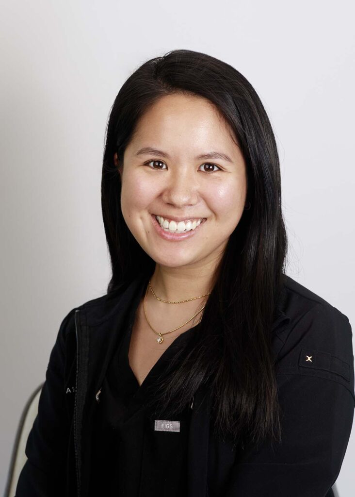 A dentist smiling with long, straight black hair wearing a black jacket with the text 'Figs' on the right side.