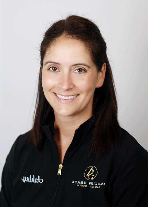 The image shows a woman smiling with a straight brown hair and wearing a black jacket.