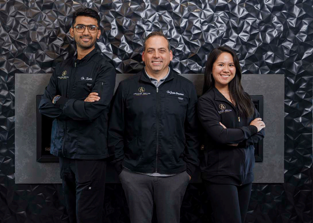 The image shows three people standing in front of a textured wall. They are all wearing black jackets with logos and text on them. The man on the left is wearing glasses and has their arms crossed. The man in the middle has their hands in their pockets, and the woman on the right also has their arms crossed.