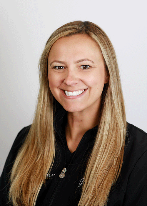 The image shows a woman smiling with a long straight blonde hair and wearing a black jacket.
