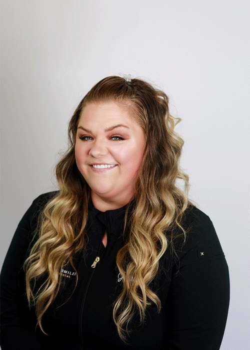 The image shows a woman smiling with a long medium curls blonde hair and wearing a black jacket.