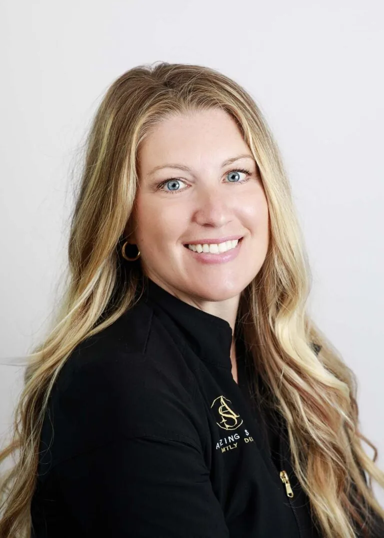 A dentist smiling with long, wavy blonde hair wearing a black jacket with the text 'AMAZING SMILES FAMILY DENTAL' on the left side.