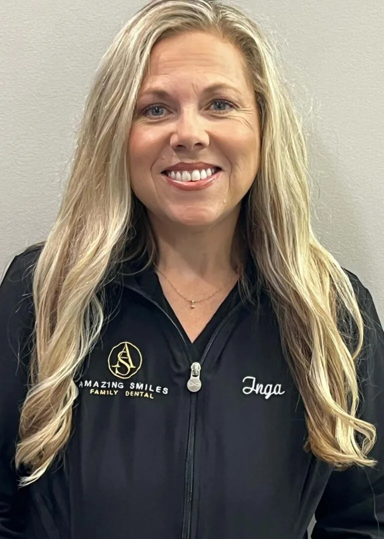 A dentist smiling with long, wavy blonde hair wearing a black jacket with the text 'AMAZING SMILES FAMILY DENTAL' on the left side and the name 'Inga' on the right.