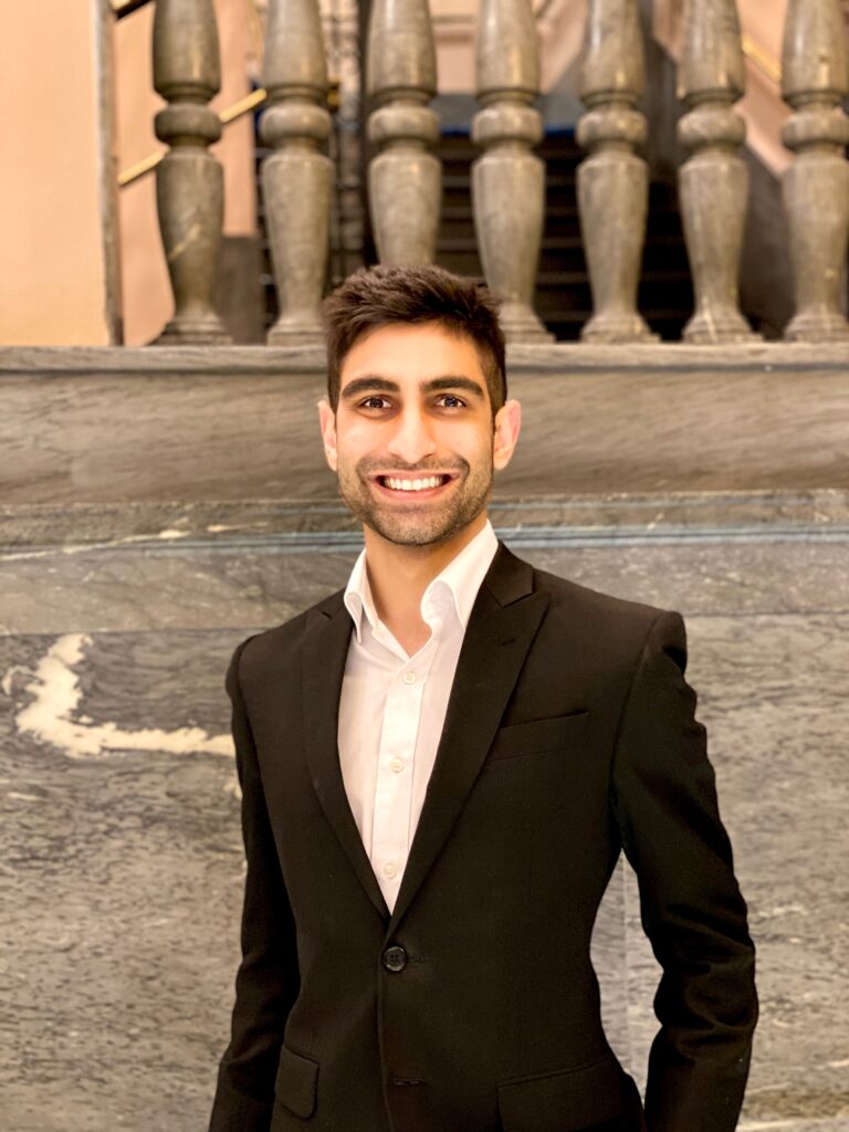 A person in a black suit and white dress shirt stands confidently in front of a marble wall, with a grand staircase featuring ornate balusters behind them. The setting suggests an elegant or historic venue, possibly for a formal event.