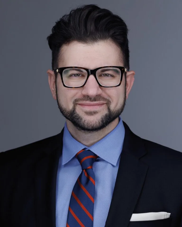 Dr. Saeed Gholizadeh in formal attire wears a dark suit jacket with a white pocket square, a light blue dress shirt, and a blue-and-red striped tie, standing against a plain background.