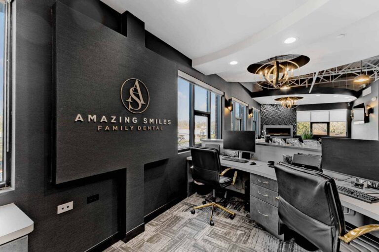 The image shows the interior of "Amazing Smiles Family Dental," featuring a sleek design with dark walls, large windows, and stylish lighting. There are multiple workstations with computers and office chairs. The dental office's logo and name are displayed on a wall to the left.