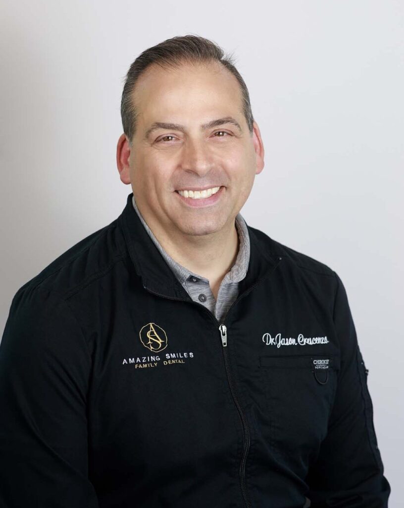 The image shows a dentist smiling wearing a black jacket with two logos and text on it. The left side of the jacket has a logo with the text "AMAZING SMILES FAMILY DENTAL." The right side of the jacket has the text "Dr. Jason Crescenzo."