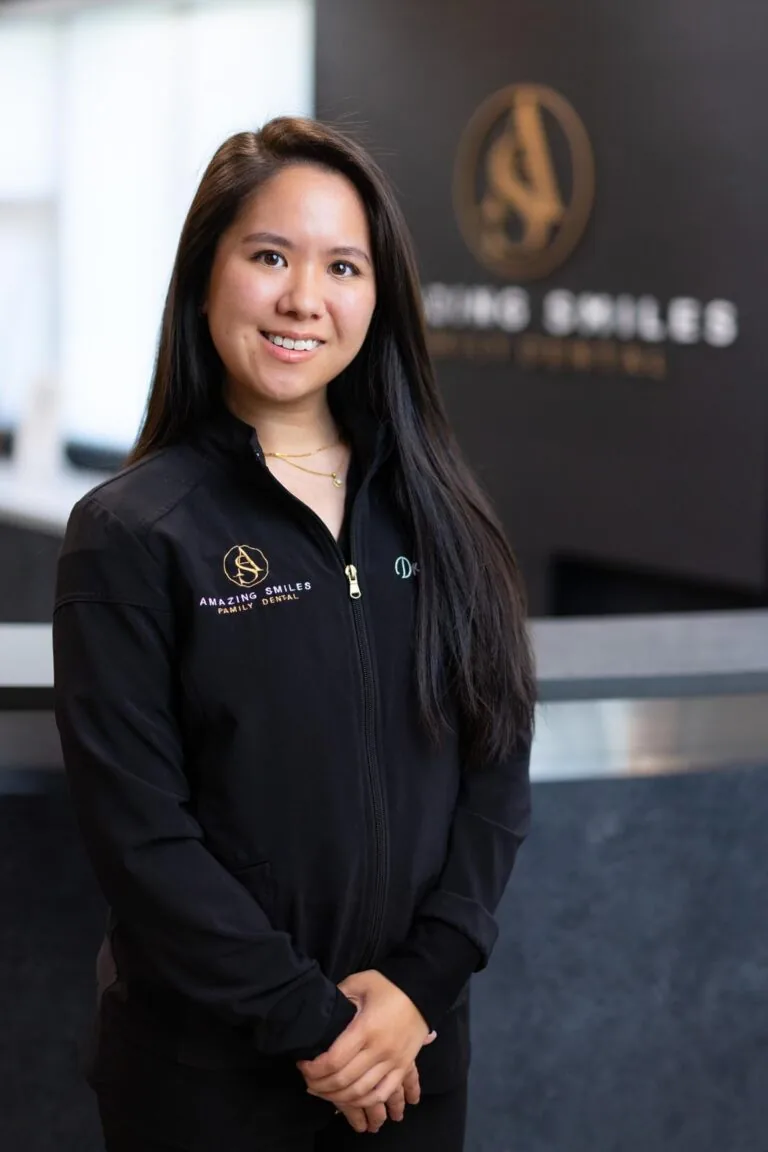 Dr. Emily Hoang headshot at Amazing Smiles lobby. Stands indoors in front of a reception desk wearing a black jacket embroidered with “AMAZING SMILES.”