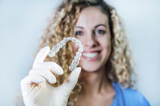 A person wearing a blue shirt and white gloves is holding a clear dental aligner in focus. The clearly visible, showing the detailed structure of the teeth. The background is plain and out of focus, emphasizing the aligner.