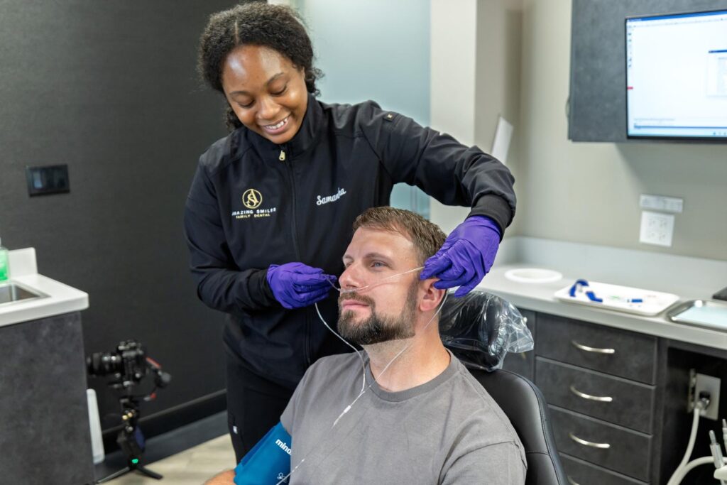 Samantha from "Amazing Smiles" is putting a nasal cannula on a male patient with a beard. The patient is sitting in a dental chair, and his arm is wrapped with a blood pressure cuff.