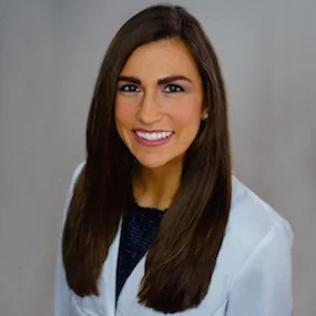 The image shows a person smiling in a white lab coat over a dark top, standing against a plain, light-colored background.