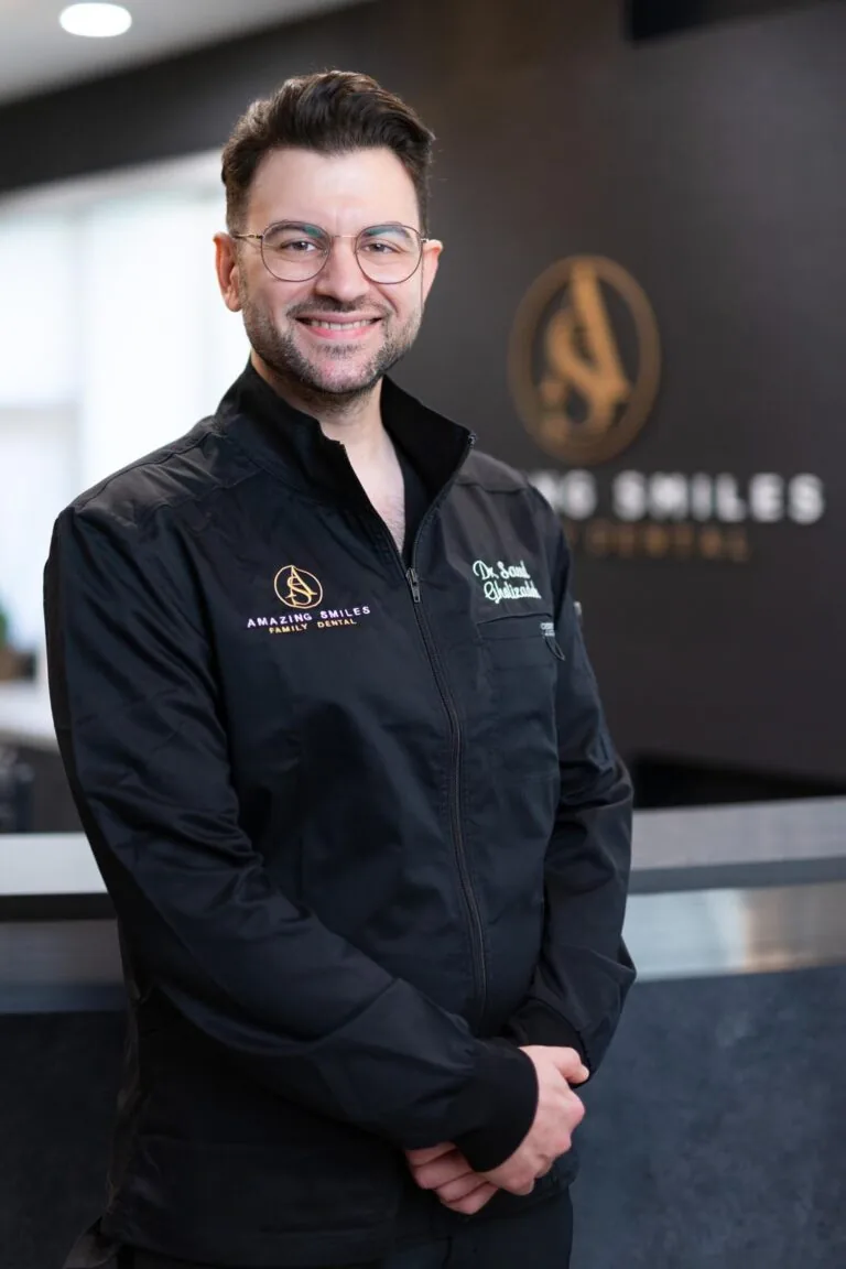 Dr. Saeed Gholizadeh headshot at Amazing Smiles lobby. Stands indoors in front of a reception desk wearing a black jacket embroidered with “AMAZING SMILES.”