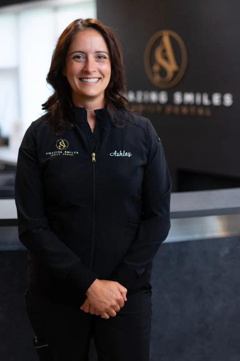 Ashley Hygienist headshot at Amazing Smiles lobby. Stands indoors in front of a reception desk wearing a black jacket embroidered with “AMAZING SMILES.”