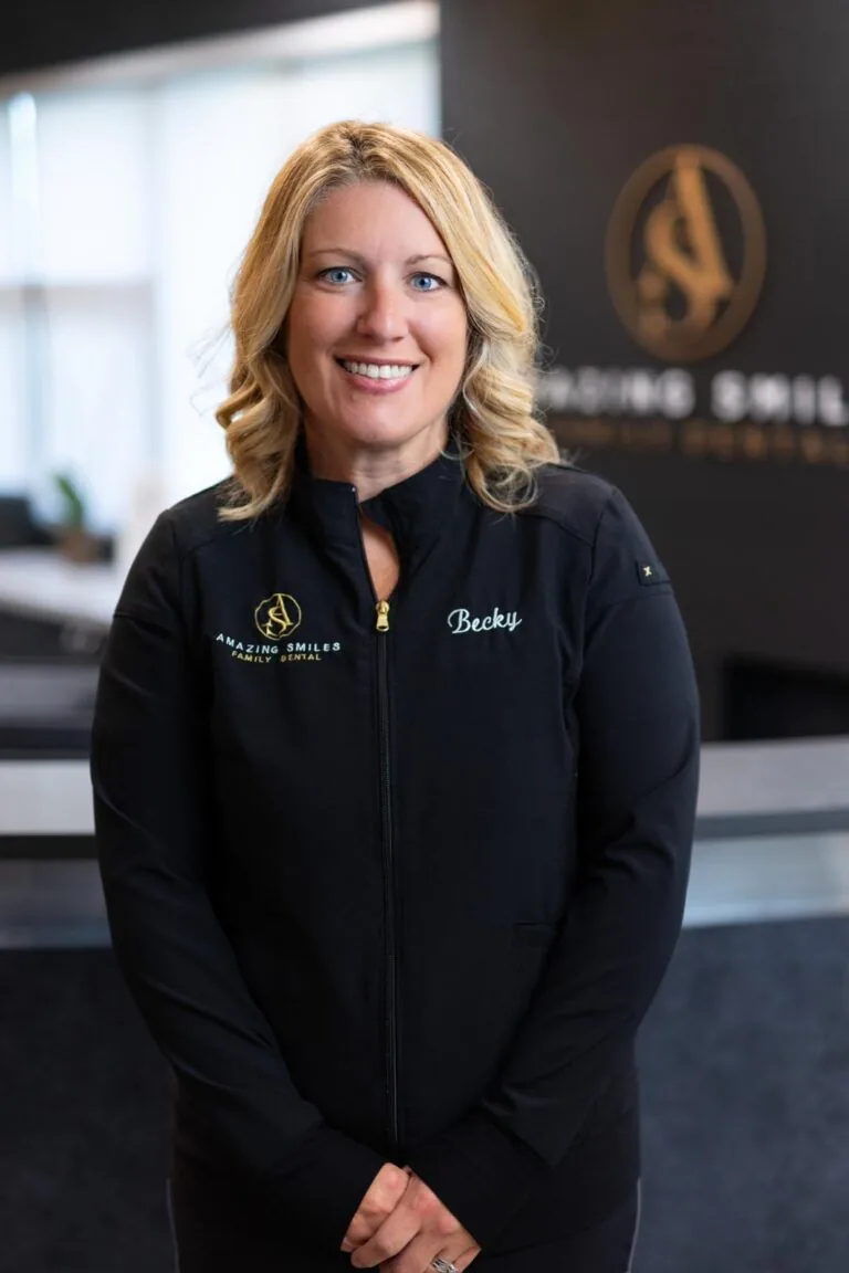 Becky Lead Hygienist headshot at Amazing Smiles lobby. Stands indoors in front of a reception desk wearing a black jacket embroidered with “AMAZING SMILES.”