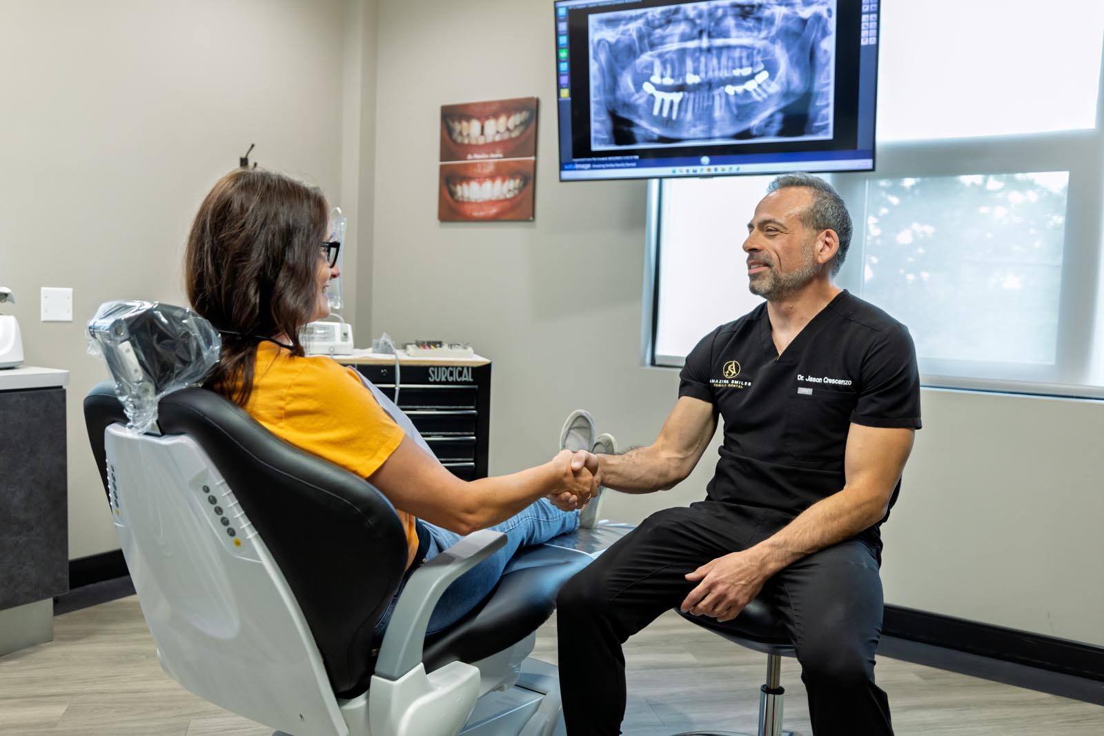 Dental consultation scene with a patient seated in a chair shaking hands with a dentist in black scrubs. Behind them is a monitor showing a panoramic dental X-ray and a cabinet marked “SURGICAL.”
