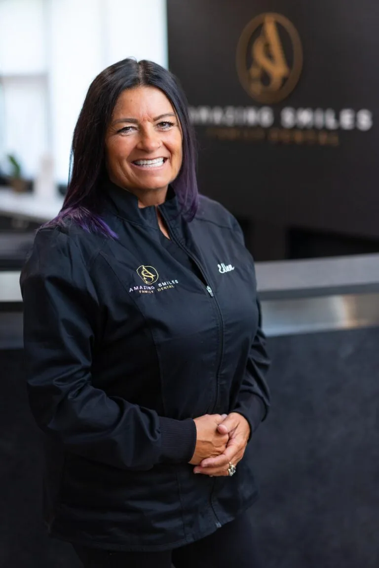 Ellen Marketing and Insurance Lead headshot at Amazing Smiles lobby. Stands indoors in front of a reception desk wearing a black jacket embroidered with “AMAZING SMILES.”