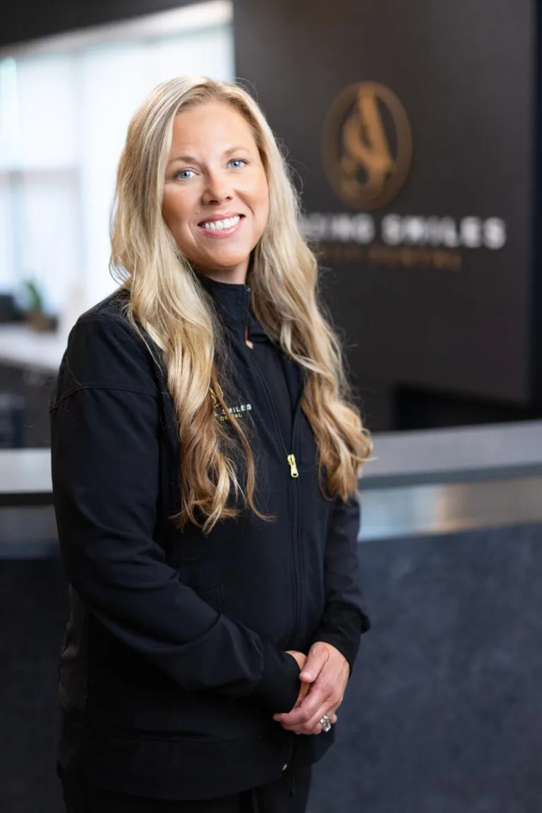 Inga S Hygienist headshot at Amazing Smiles lobby. Stands indoors in front of a reception desk wearing a black jacket embroidered with “AMAZING SMILES.”