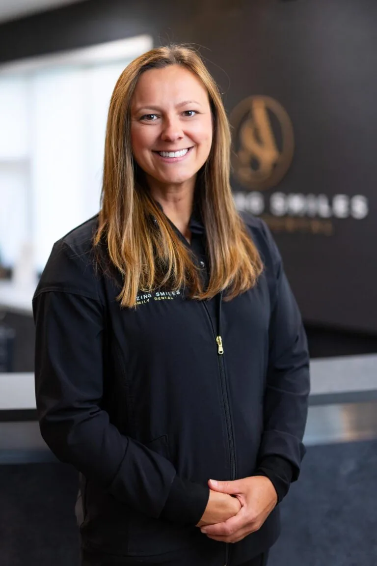 JJ Hygienist headshot at Amazing Smiles lobby. Stands indoors in front of a reception desk wearing a black jacket embroidered with “AMAZING SMILES.”