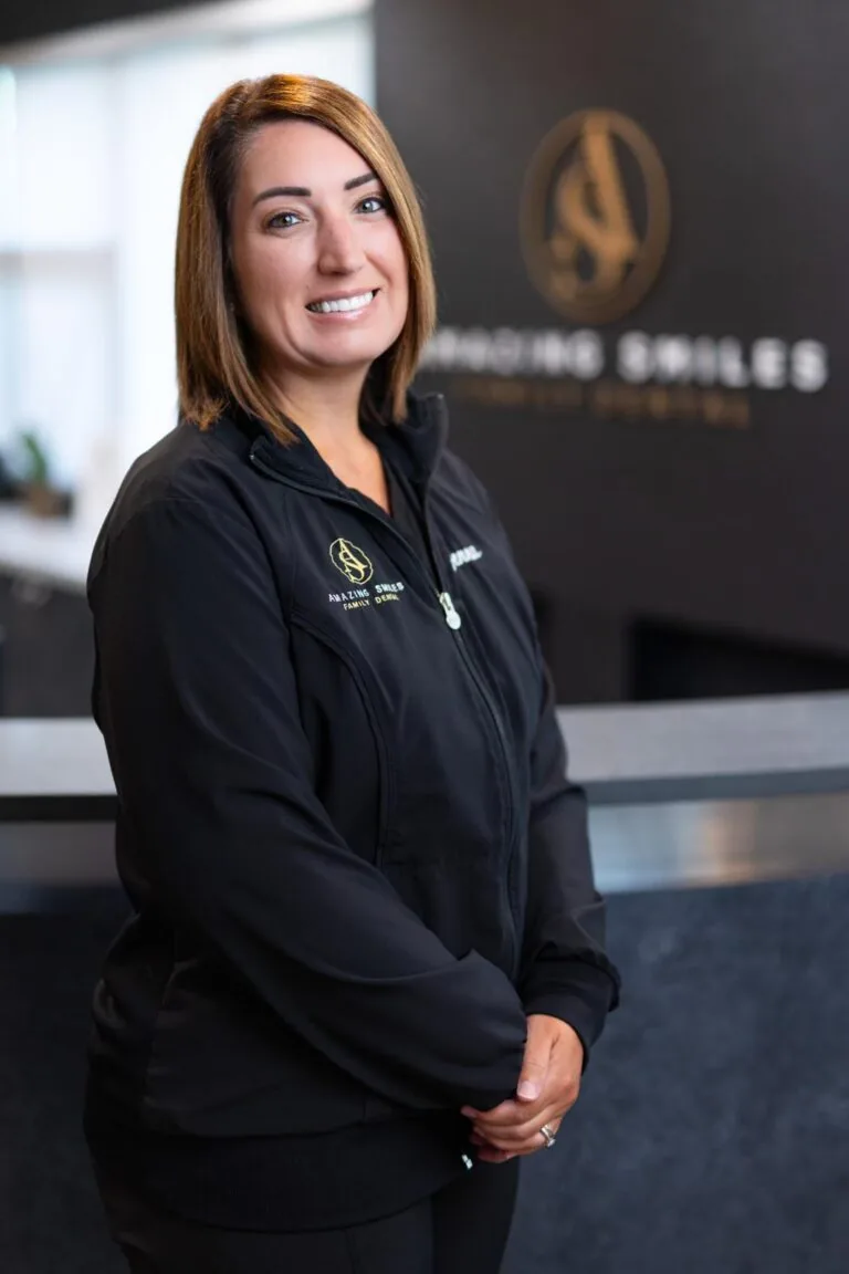 Jenna Hygiene Lead headshot at Amazing Smiles lobby. Stands indoors in front of a reception desk wearing a black jacket embroidered with “AMAZING SMILES.”