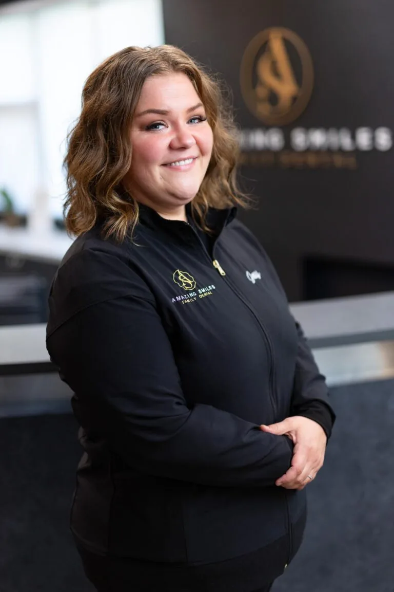 Jessy Hygienist headshot at Amazing Smiles lobby. Stands indoors in front of a reception desk wearing a black jacket embroidered with “AMAZING SMILES.”