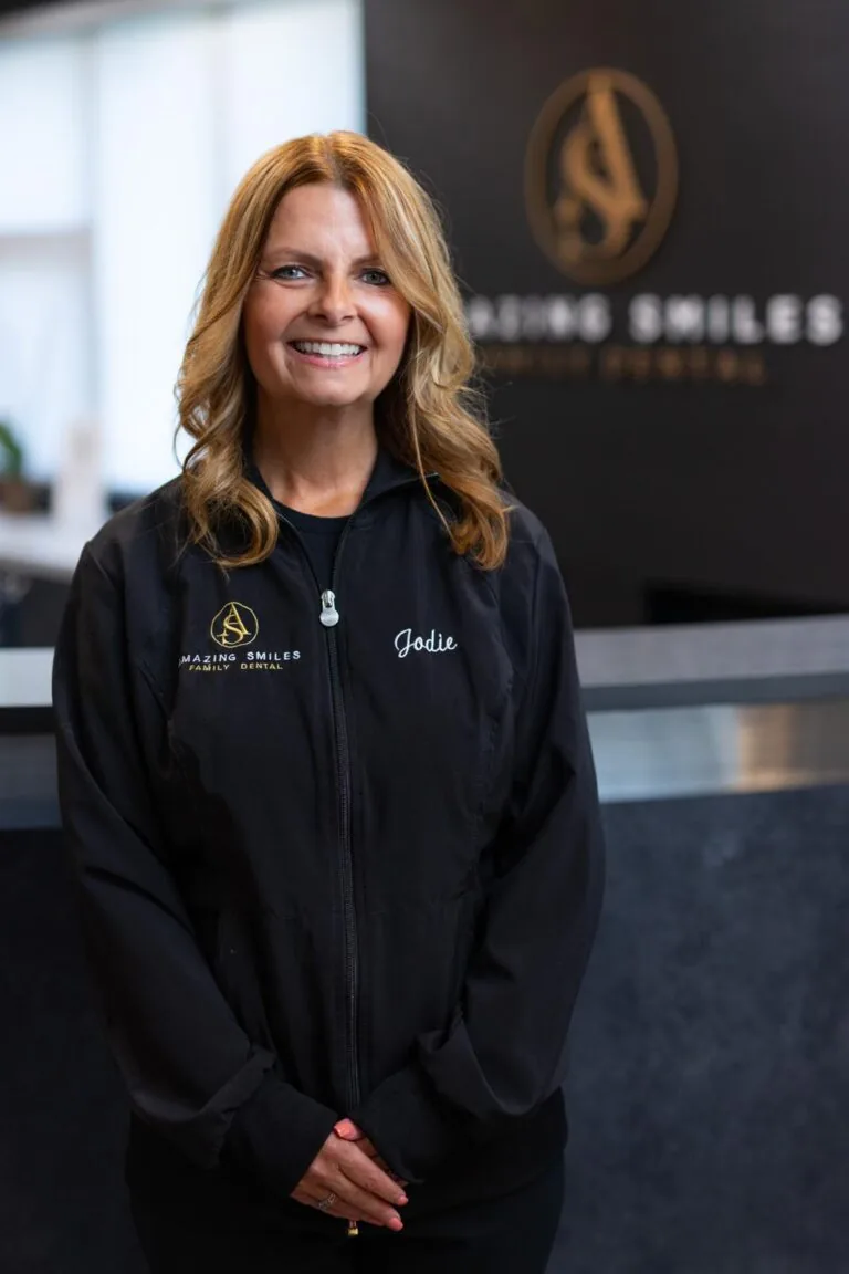 Jodie Office Manager headshot at Amazing Smiles lobby. Stands indoors in front of a reception desk wearing a black jacket embroidered with “AMAZING SMILES.”