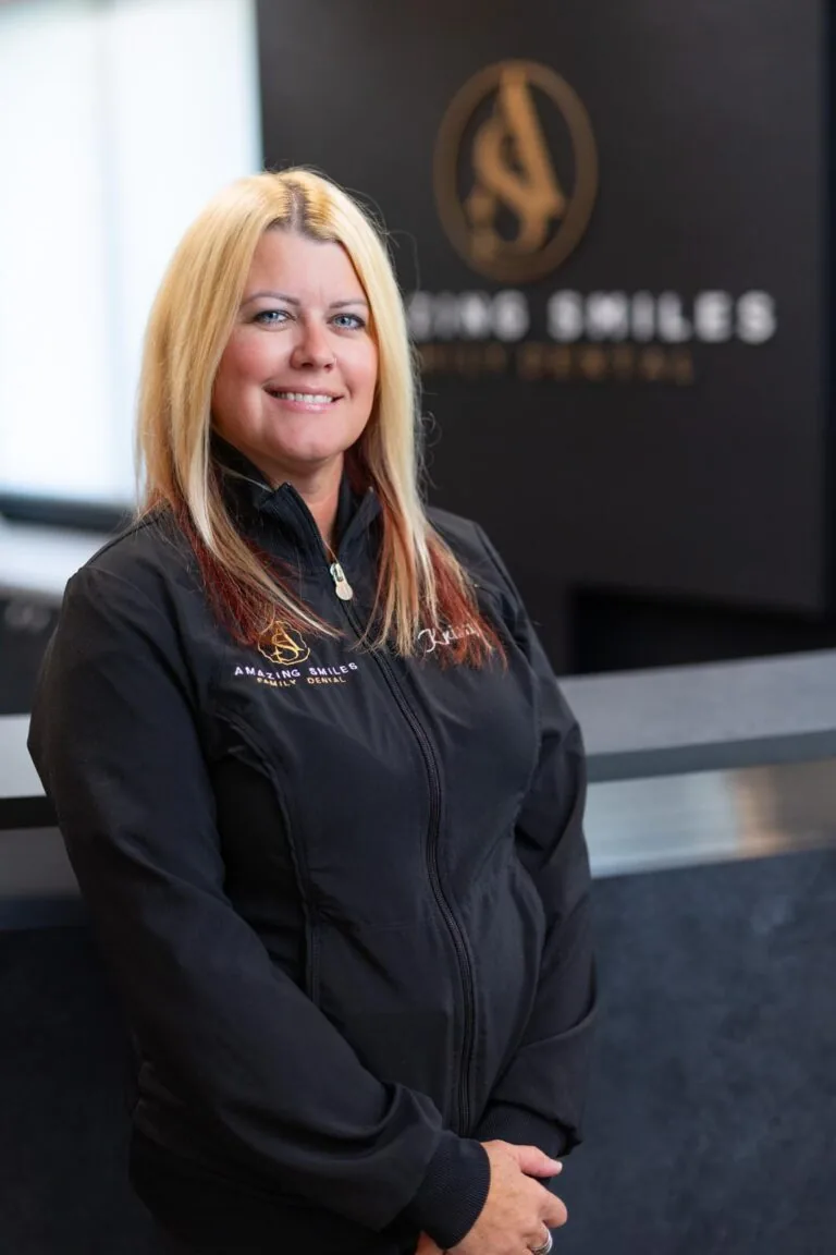 Kristy Treatment Coordinator Lead headshot at Amazing Smiles lobby. Stands indoors in front of a reception desk wearing a black jacket embroidered with “AMAZING SMILES.”