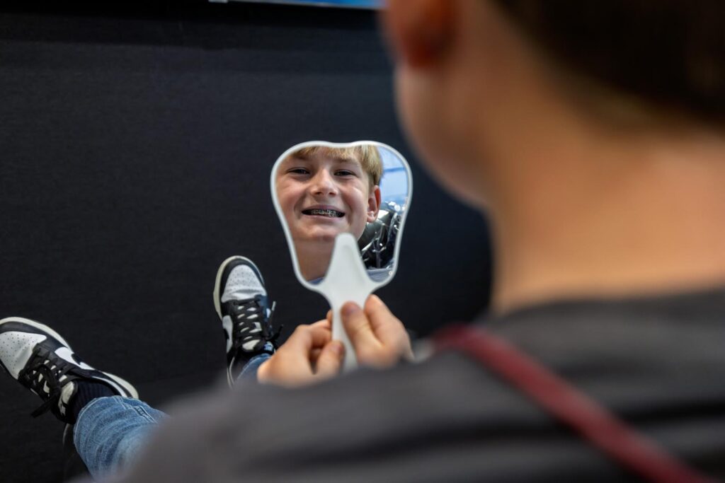 A patient holds a tooth-shaped mirror, reflecting the smiling face of a young boy with braces on his teeth. 