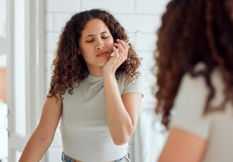 Woman with curly hair touching jaw in discomfort while holding toothbrush, showing possible TMJ pain in bathroom mirror.